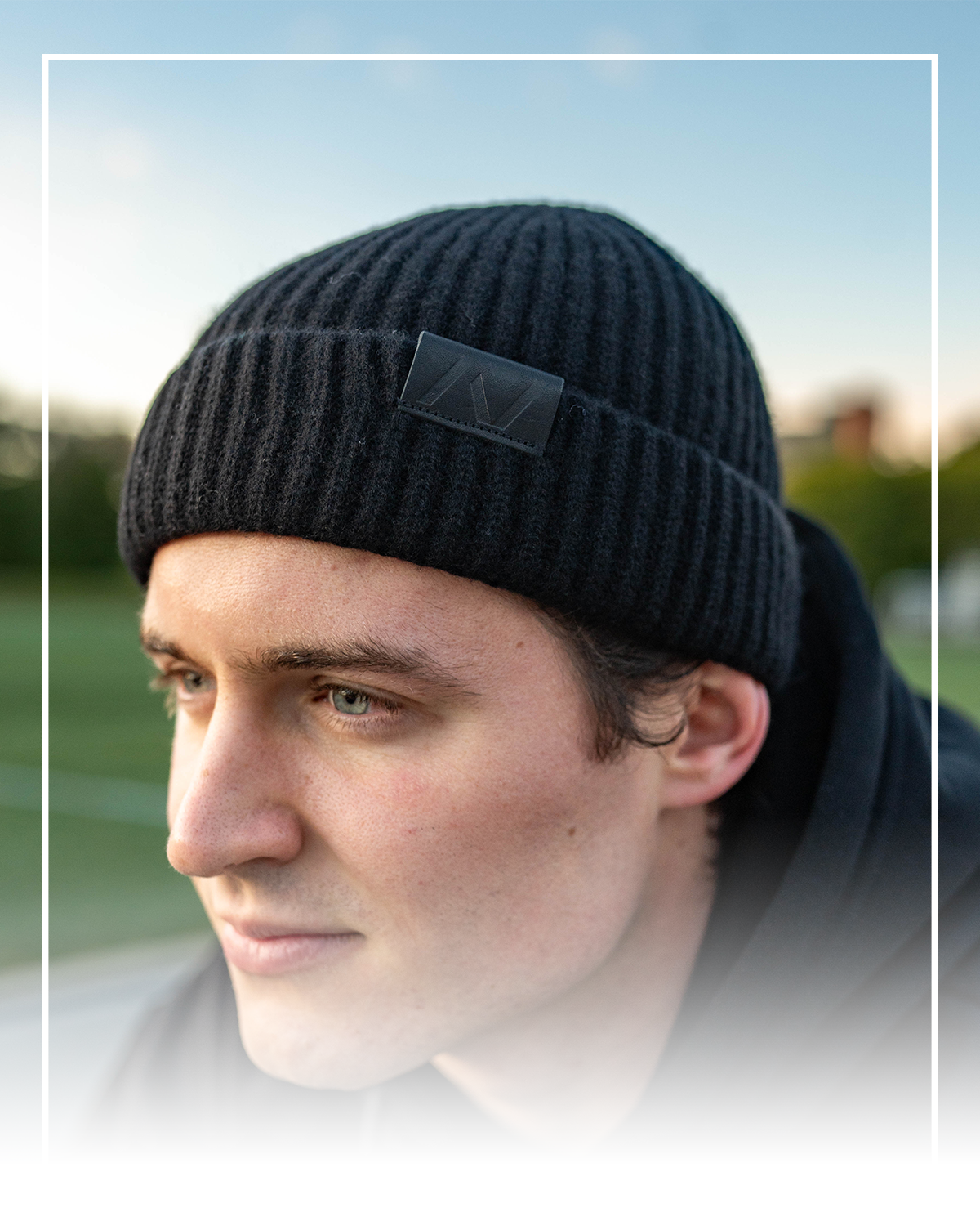 Person wearing a black beanie with a logo against a blurred outdoor background