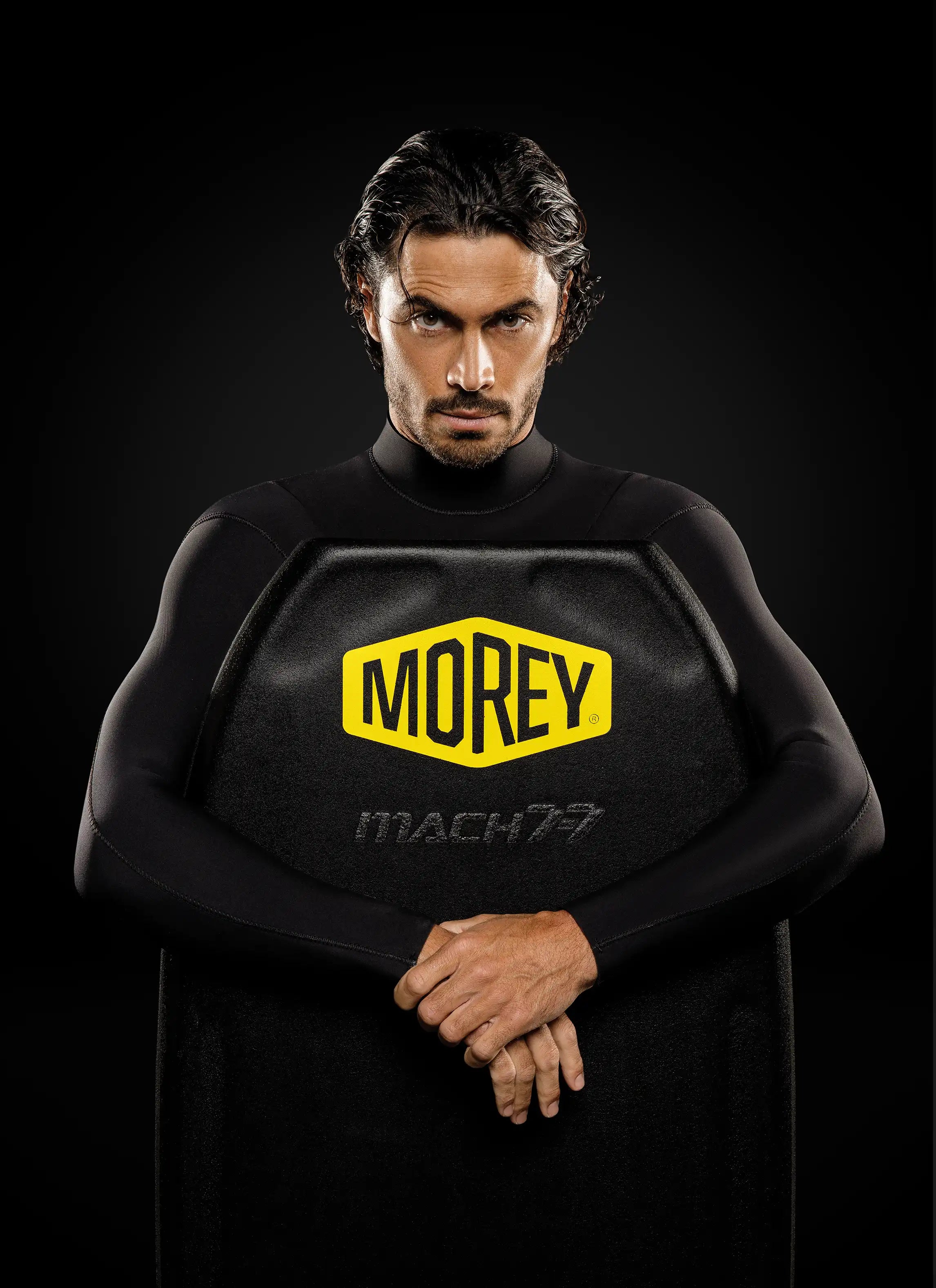 Man in wetsuit holding black Morey Mach 7 bodyboard against a dark background