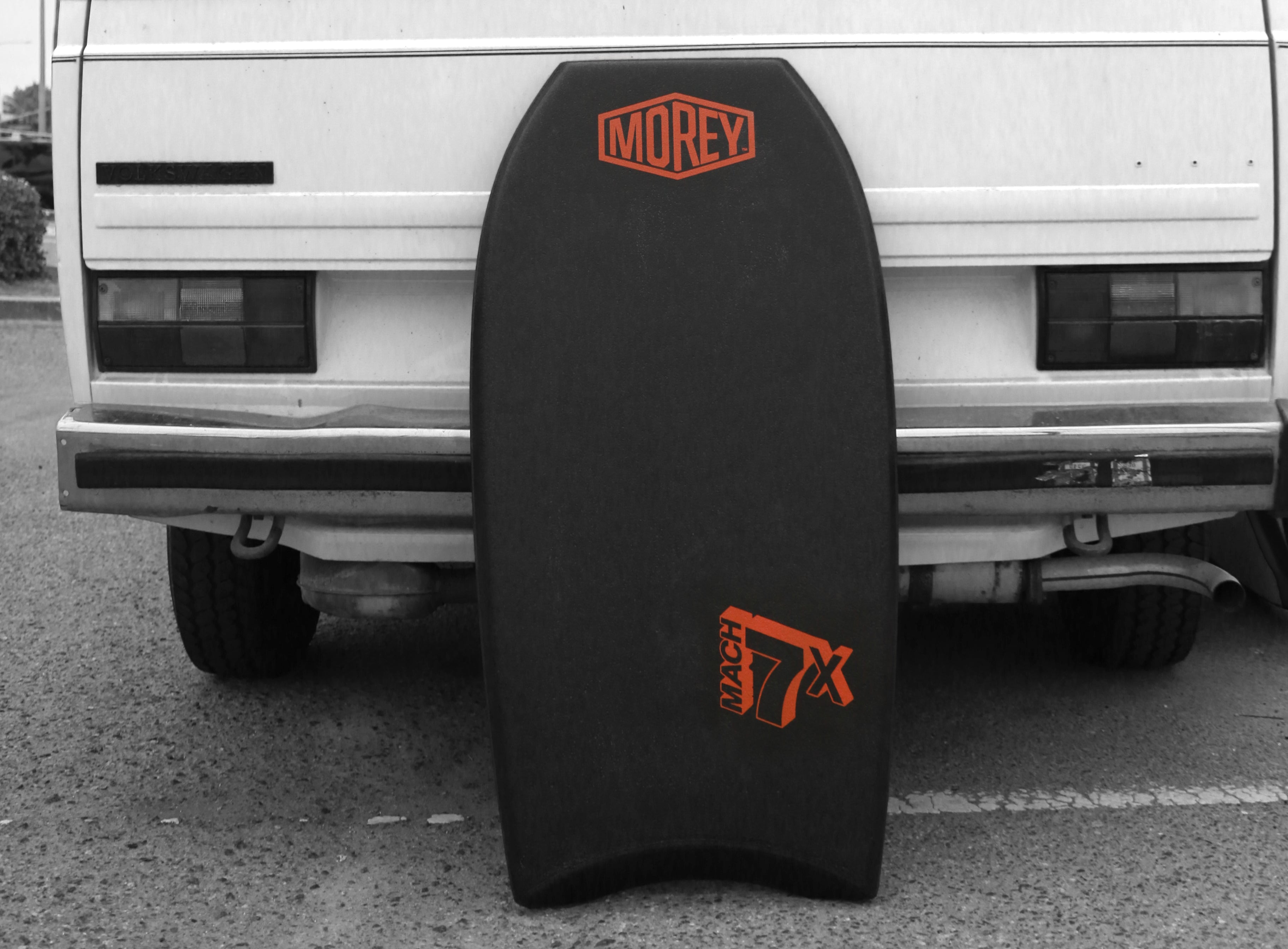 Black Mach 7X bodyboard with orange logo leaning against a white vehicle in a parking lot