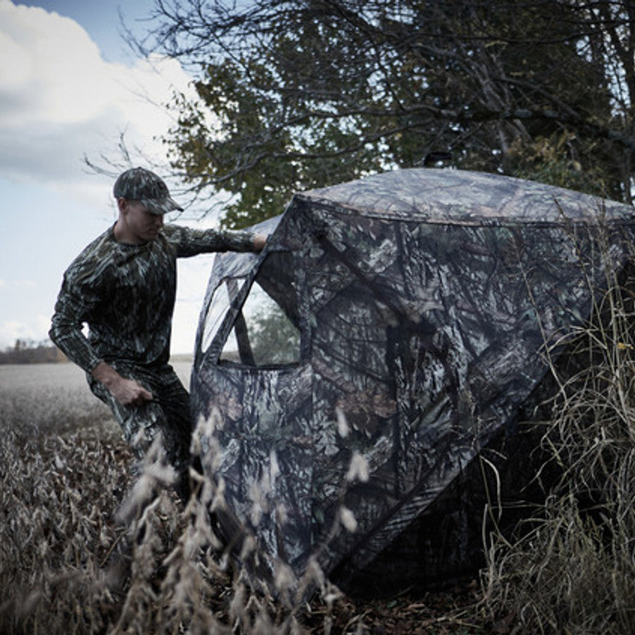 Ameristep Magnum Care Taker 59x59 pop-up ground blind in Mossy Oak camouflage with hunter setting up blind in open field near tree line