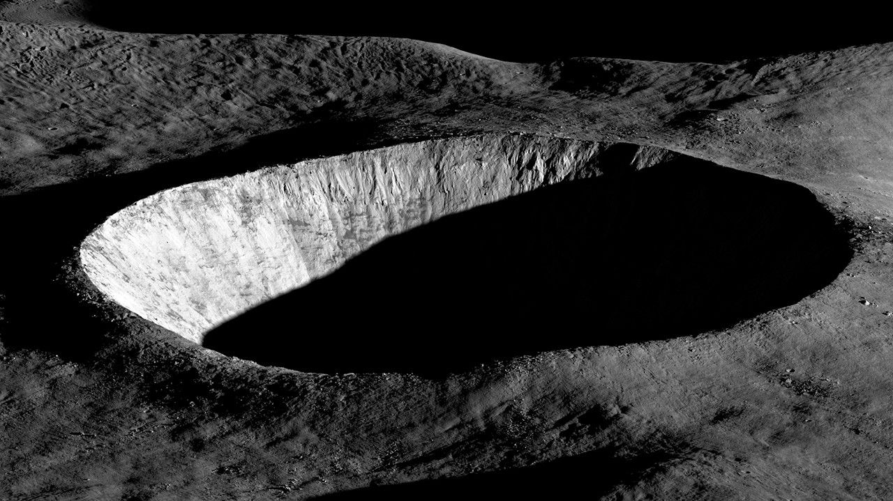 A black and white photograph of an unnamed lunar crater captured by NASA's Lunar Reconnaissance Orbiter Camera (LROC) on August 30, 2023. The image shows dramatic contrast between light and shadow as early morning sunlight illuminates only the western wall of the crater, which appears bright white against the dark lunar landscape. The interior of the crater remains in complete shadow, creating a striking bowl-shaped void. The surrounding lunar surface displays the characteristic rough, textured terrain of the Moon with various small craters and ridges visible. This high-resolution image demonstrates LROC's capability to capture detailed topographical features, part of LRO's mission to map the lunar surface for future exploration efforts.