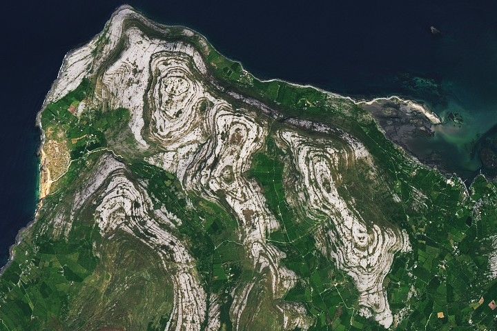 A detailed view of folded limestone in the Burren. The limestone forms swirling, layered patterns of gray rock separated by thin green bands of vegetation.