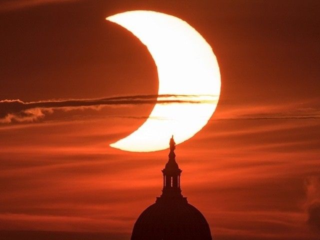 Close-up of the Sun partially blocked by the Moon over a domed, statue-topped building, with dramatic orange-lit clouds in the foreground.