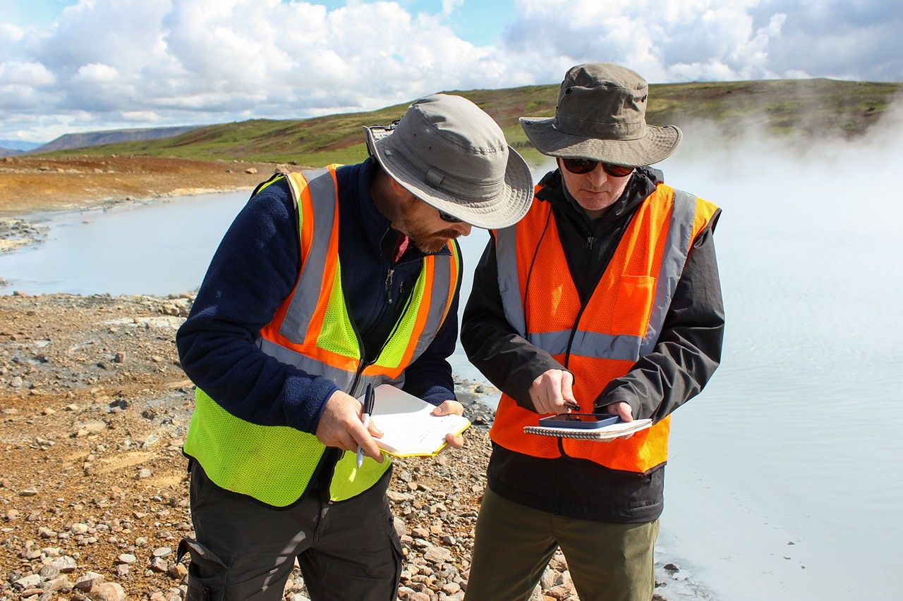 Two scientists in safety vests and sun hats examine field notes and samples beside a steaming geothermal pool with mineral-stained shores in bright sunlight.