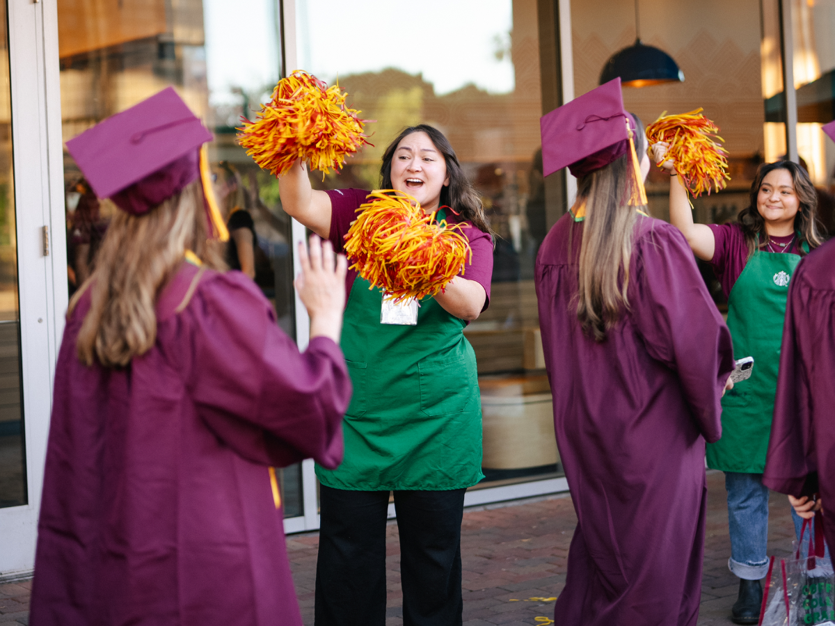 At a Starbucks College Achievement Plan graduation at Arizona State University, graduates walk past a Starbucks employee wearing a green apron who is cheering them on with pompoms.