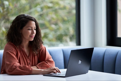Orang mengenakan blus oranye bekerja menggunakan laptop di atas meja putih dengan sofa biru dan jendela di latar belakang.