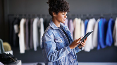 Eine junge Frau in einer Jeansjacke, die ein Tablet in einem Bekleidungsgeschäft nutzt und neben einem Kleiderständer steht.