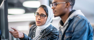 Due persone lavorano insieme al computer. La donna con il velo bianco indica lo schermo, concentrata e coinvolta nella discussione.