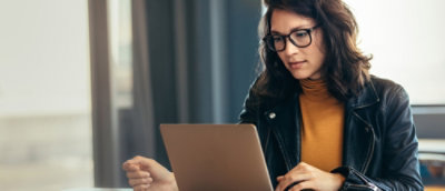 A woman with long hair wearing glasses, looking at a laptop.
