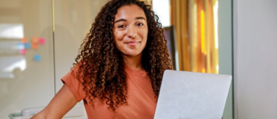 Una persona con el pelo rizado sonríe cálidamente a la cámara mientras sostiene un portátil.