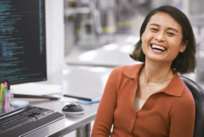 Eine lächelnde Person, die an einem Schreibtisch mit einem Computer sitzt, auf dessen Bildschirm Codezeilen angezeigt werden.