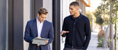 Two men standing outside, one in a suit holding a tablet, and the other looking at it.