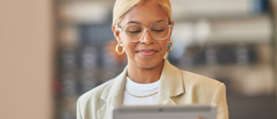Une professionnelle portant des lunettes et une veste légère beige qui lit sur sa tablette dans un bureau moderne.
