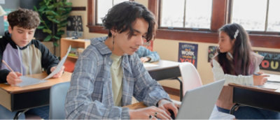 Three students in a classroom; one uses a laptop, another writes on paper, and the third looks sideways.