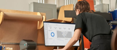 Une personne, vêtue d’une chemise noire et penchée sur son bureau, qui interagit avec des données sur un grand écran de bureau affichant des graphiques et un mappage