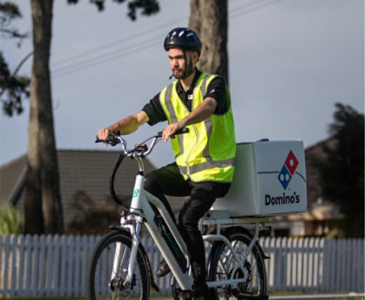 Eine Person, die Fahrrad mit einer Pizzabox fährt, die eine Sicherheitsweste und einen Helm trägt.
