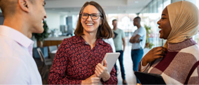Three people are having a friendly conversation in an office setting. One woman holds a tablet, and another person has a notepad.