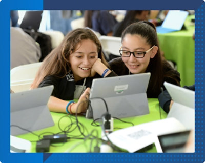 Two young girls are smiling and looking at a tablet screen together. Various electronic devices and cables are on a green table around them.