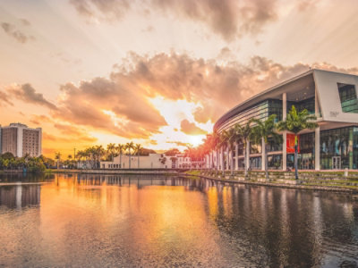 Sunset over stunning modern architecture and calm water in Miami, Florida, with vibrant clouds and palm trees reflecting in the lake.