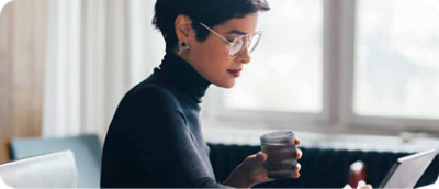 Kvinne med kort hår og briller holder et krus mens hun ser på en bærbar datamaskin i et lyst rom.