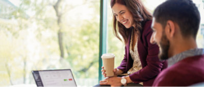 Two people sit together, one holding a cup, smiling and looking at a laptop screen. Bright window view in the background.