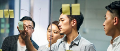 A group of four professionals collaborating in a modern office setting, with one person writing on a glass board while others observe and engage in discussion.