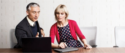 Een man en een vrouw zitten aan een tafel, bezig met een laptop voor zich, en bespreken of werken samen aan een project.