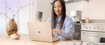 Una persona con el pelo largo sostiene una taza mientras utiliza un portátil en una mesa. Al lado, hay un bol de fruta y un jarrón.