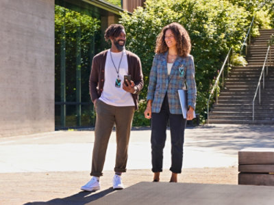 A man and a woman standing outside, with the woman wearing a plaid jacket and the man holding a phone.