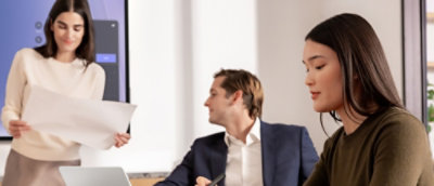 A group of people sitting at a table with a woman holding a piece of paper and a man in a suit nearby.