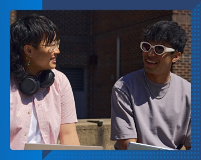 晴れた日にレンガ造りの建物の前で、互いに向かって微笑んでいる 2 人。一人はヘッドフォンと眼鏡を、もう一人はサングラスを着用しています。