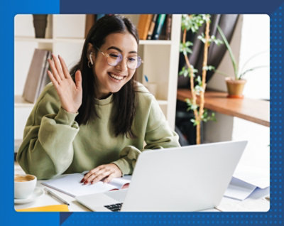 Жінка сидить перед ноутбуком і махає комусь рукою, поруч стоїть рослина в горщику.