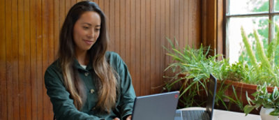 Wanita tersenyum sambil menggunakan laptop di ruangan dengan tanaman dan panel kayu.