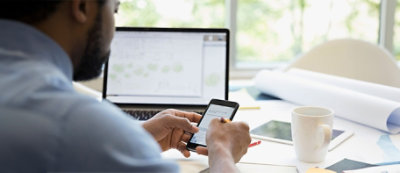 A person is using a smartphone while sitting at a desk with a laptop, tablet, cup, papers, and a pen.