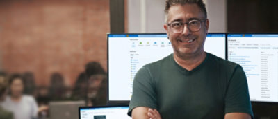 Um homem em pé em frente a dois grandes ecrãs de ambiente de trabalho e a sorrir.