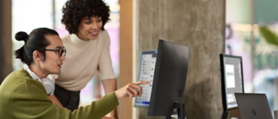 眼鏡をかけて緑色のセーターを着ている男性が見守る中、白いシャツを着た女性がコンピューターの画面を指し示しています。