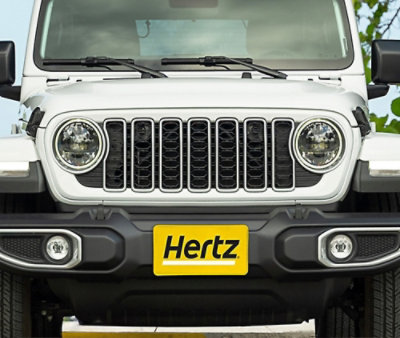 Valkoisen SUV:n etuosanäkymä, jossa on pyöreät ajovalot ja säleikkö sekä keltainen Hertz-rekisterikilpi.