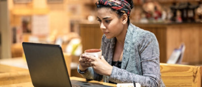 Person seated at a café table, holding a cup while working on a laptop, with a warm, casual indoor setting in the background.