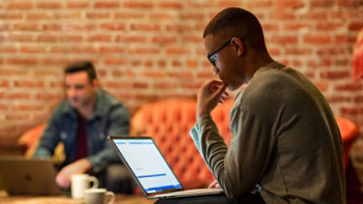 Person working on a laptop at a table in a casual office lounge with brick walls, seating, and another workspace visible in the background.