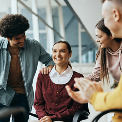 A woman in a wheelchair is congratulated by several colleagues in a modern office.