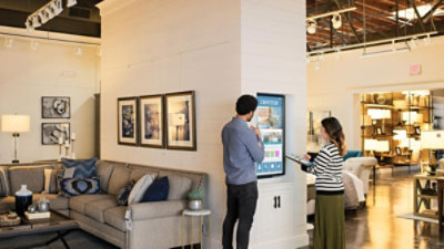 Un empleado y un cliente en una tienda de muebles observando un cartel digital con información sobre productos