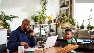 A person with an open laptop talks on the phone while his child uses a tablet