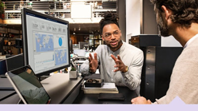 Two colleagues in a modern office discuss analytics displayed on a large monitor with maps, charts, and metrics.