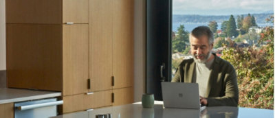 Un hombre trabajando en un portátil en un escritorio de cocina con una vista de lago en el fondo.