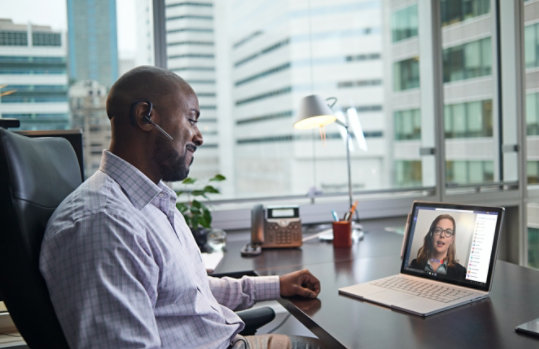 Человек сидит за столом в офисе, а на экране ноутбука отображается вызов Teams