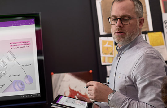 Фотография человека в профессиональной творческой студии, который работает на планшете с помощью пера. Проект отображается на этом планшете, а также на большом мониторе. 