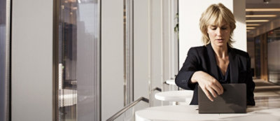 Osoba ubrana w strój biznesowy siedzi przy oknie w nowoczesnym biurze, kładąc otwartego laptopa na stole przed sobą.