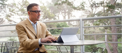 A man in a tan blazer works on a tablet at an outdoor table on a patio, surrounded by trees.