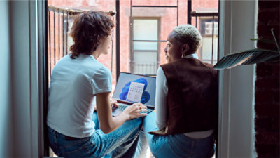Two Gen Z friends sitting on fire escape balcony with laptop open with Windows 11 start screen and start menu