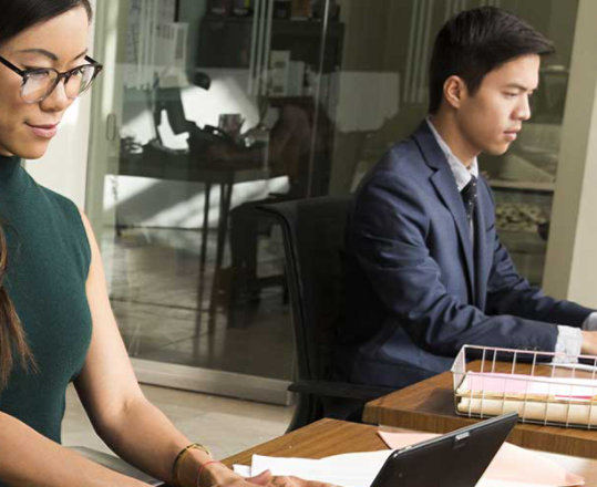 Two people working in an office on laptops with a large rack of hanging files on the table behind them.